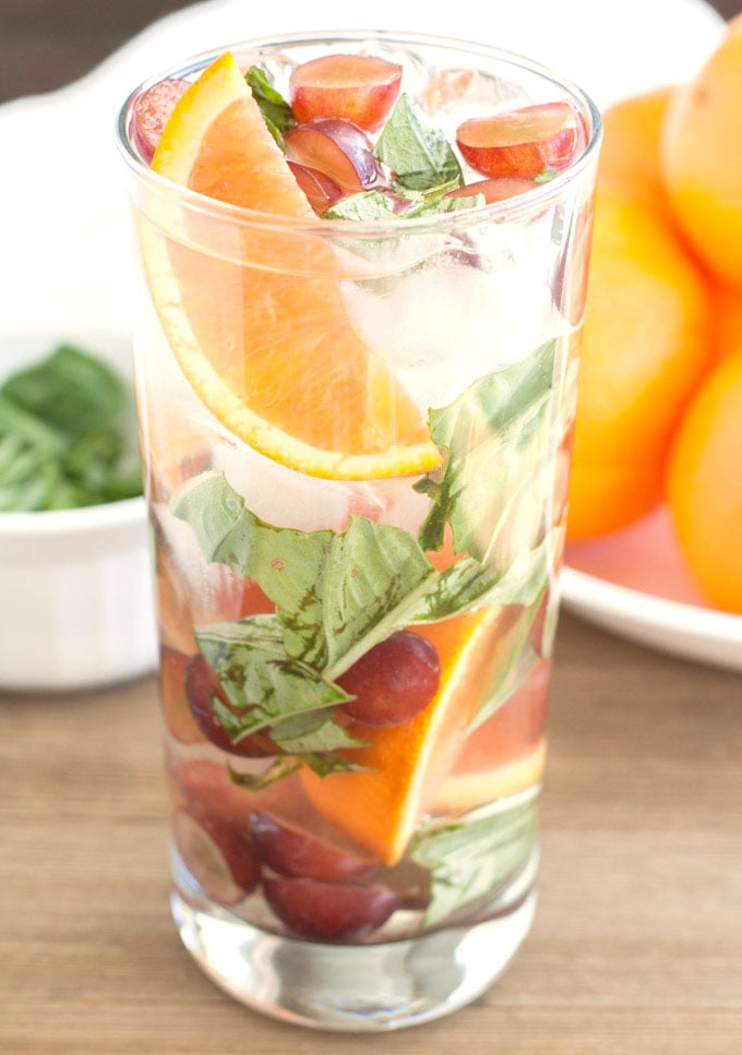 A tall glass of water with oranges, grapes and basil