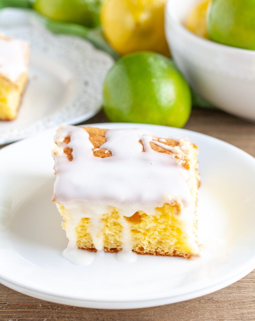 A piece cake on a plate with icing drizzling over the side and lime and lemon in background.