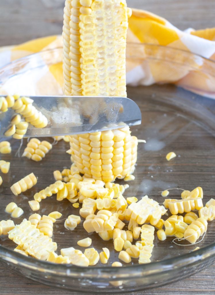 Ear of corn standing in a pie plate with knife cutting off corn kernels 