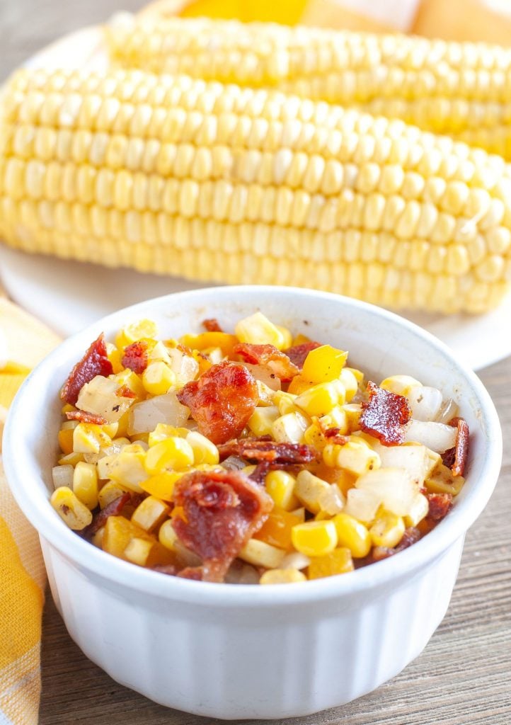 Fried corn with bacon and onions in a bowl with ears of corn in the back