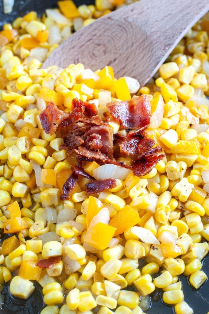 Fried Corn in a skillet topped with crumbled bacon and a wooden spoon. 