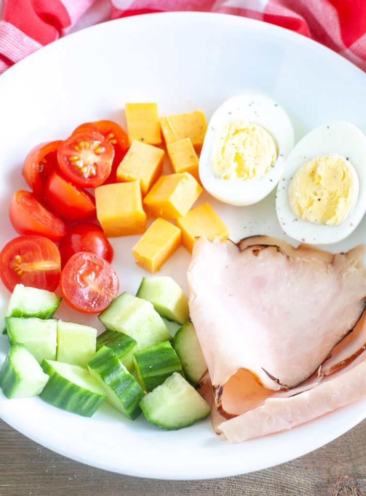 A white bowl with tomato, cheese, hard boiled egg, cucumber and turkey