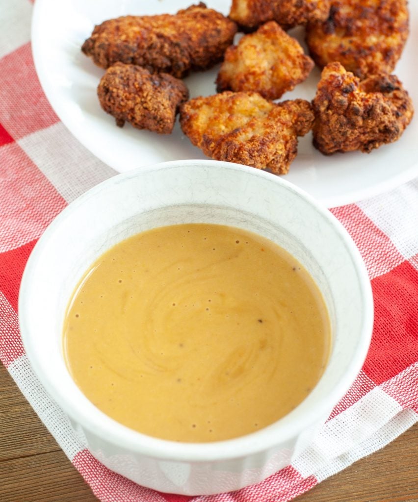 White bowl with dipping sauce and a plate with chicken nuggets on a red checked cloth