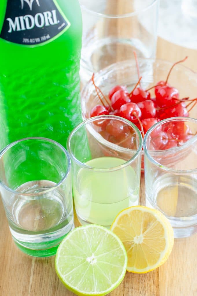bottle of midori, bowl of cherries, lemon, lime and glass with vodka