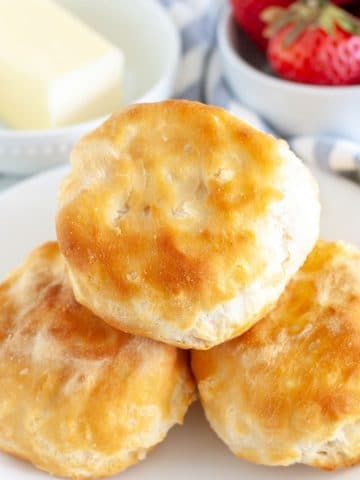 Three biscuits on a plate with bowl of butter and strawberries.