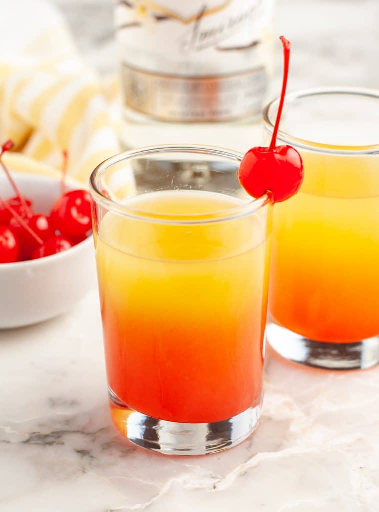 Shot glass filled with orange and red juice and a cherry.
