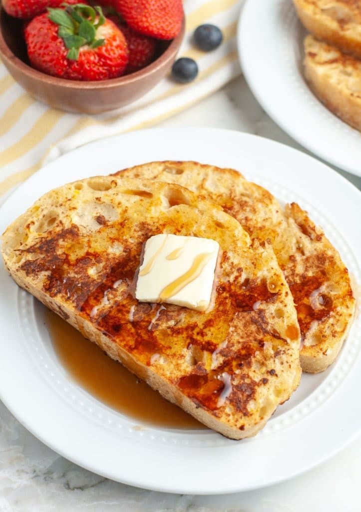 Two pieces of French toast on a plate with bowl of strawberries.