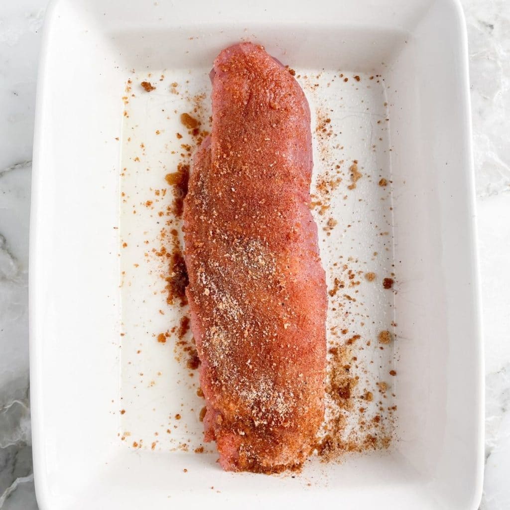 Uncooked pork tenderloin in baking dish covered in spices.