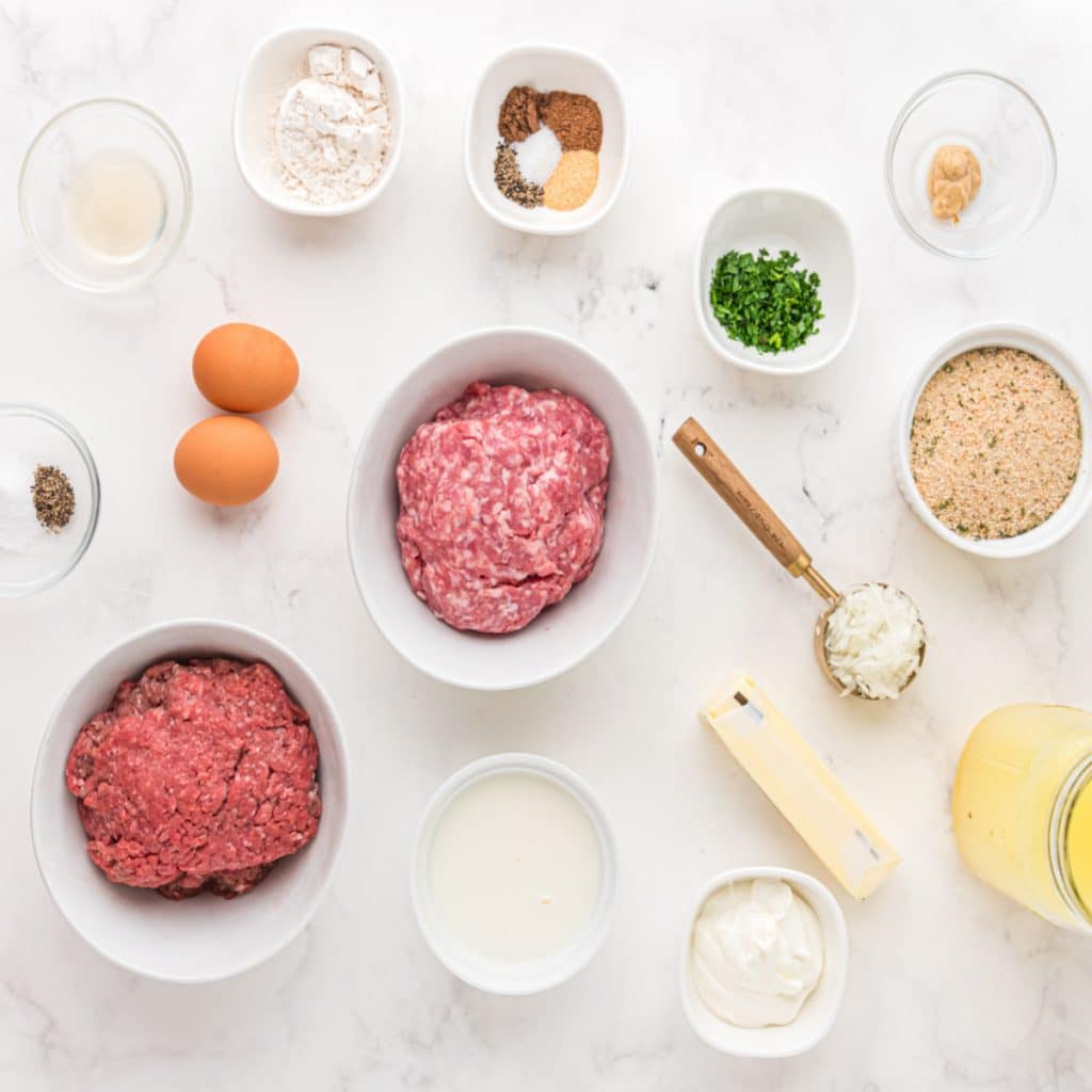 Bowls of ground beef and pork, bowl of seasoning, eggs, and bread crumbs. Stick of butter, cheese, and bowl of diced onions. 