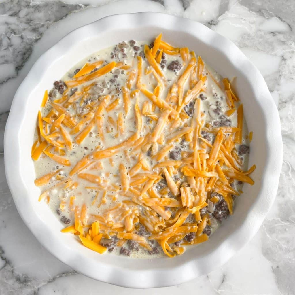 Pie plate with shredded cheese, egg mixture, and ground beef. 