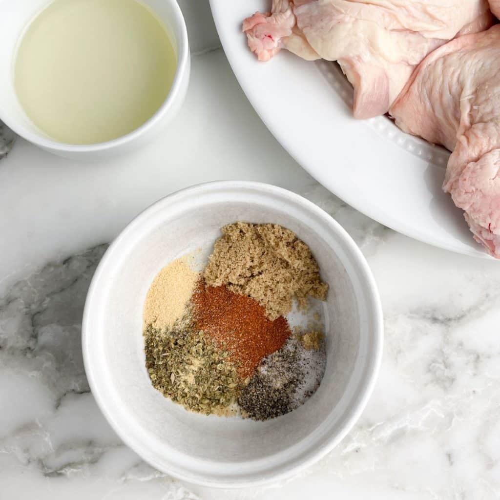 Bowl of seasonings, chicken thighs on plate. 