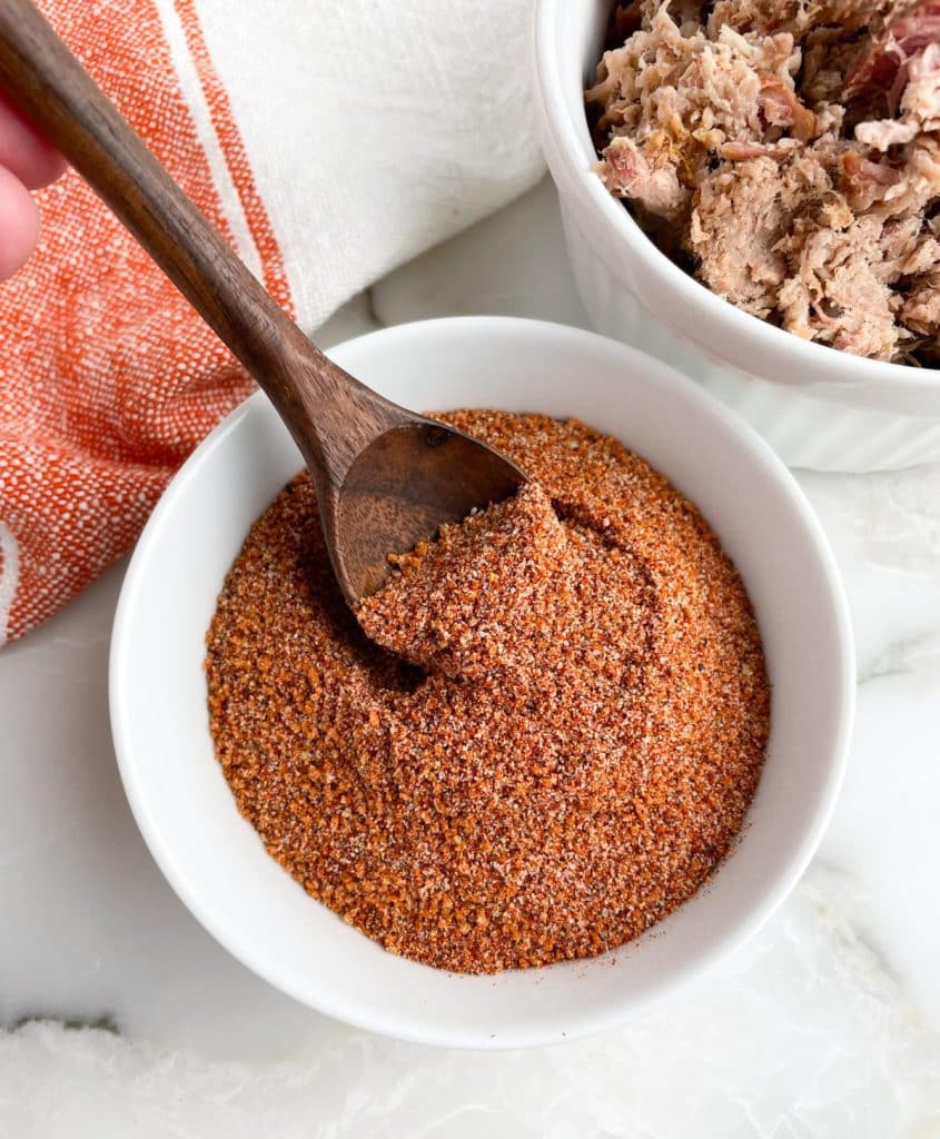 Bowl of spice mixture with a wooden spoon and bowl of pulled pork.