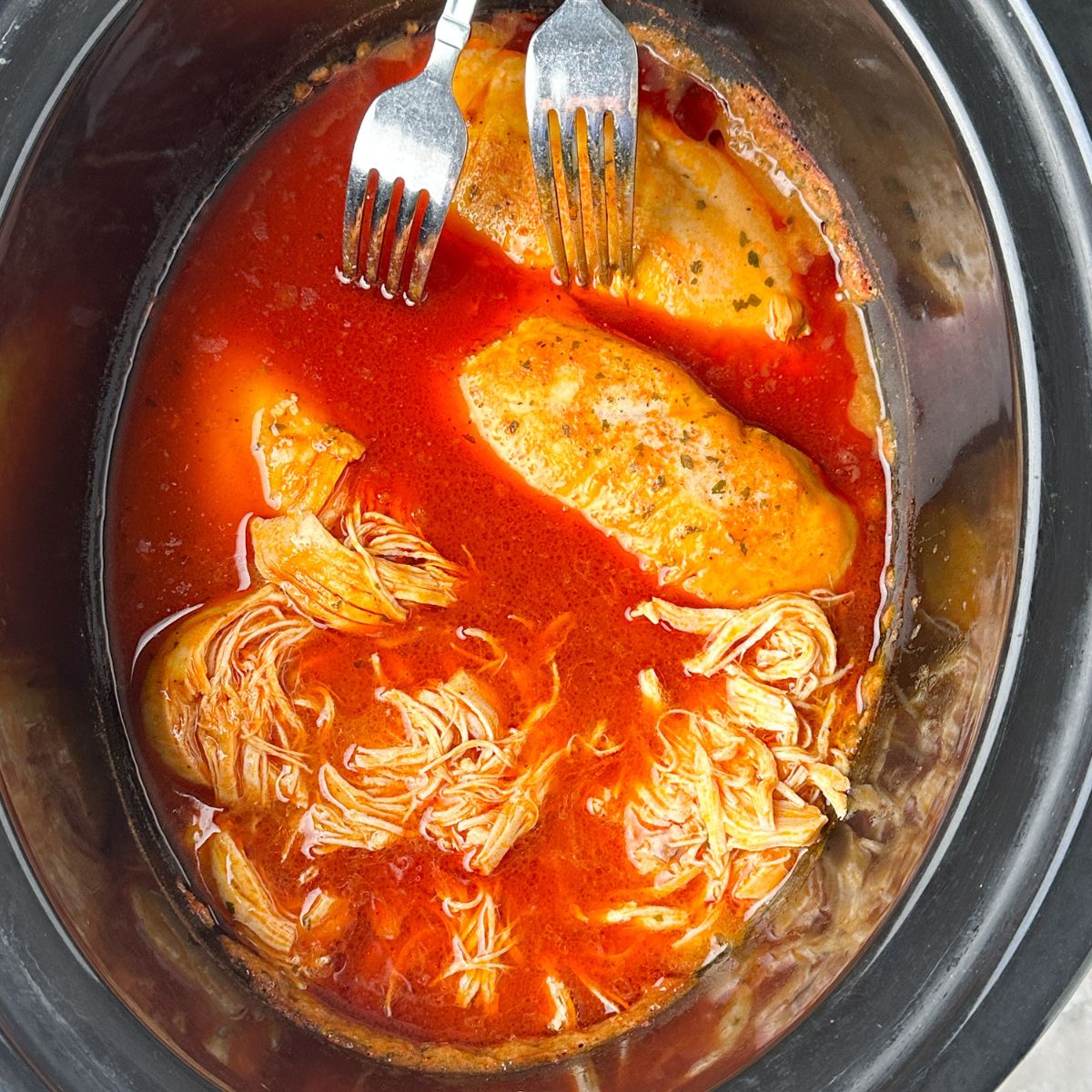 Slow cooker with chicken, buffalo sauce, and forks.