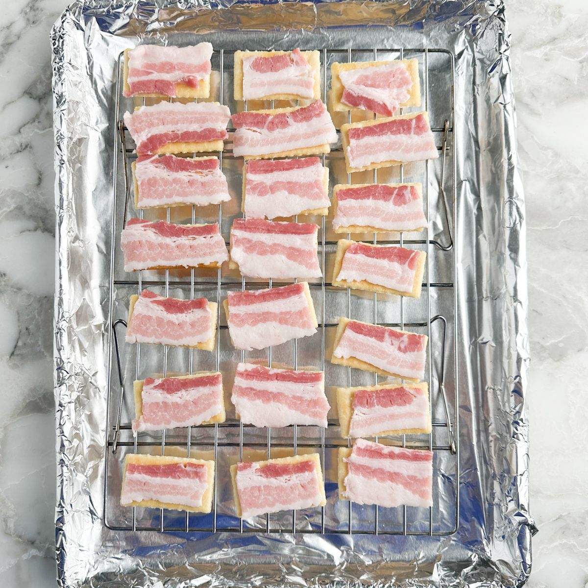 Baking tray with crackers topped with sliced of uncooked bacon. 