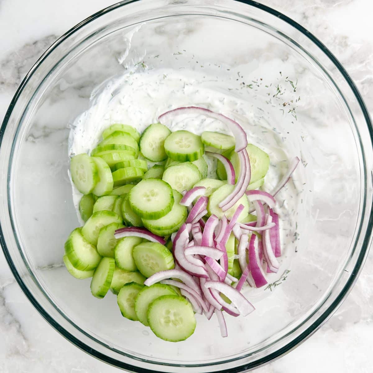 Bowl of creamy dressing, sliced cucumbers, and sliced red onion.