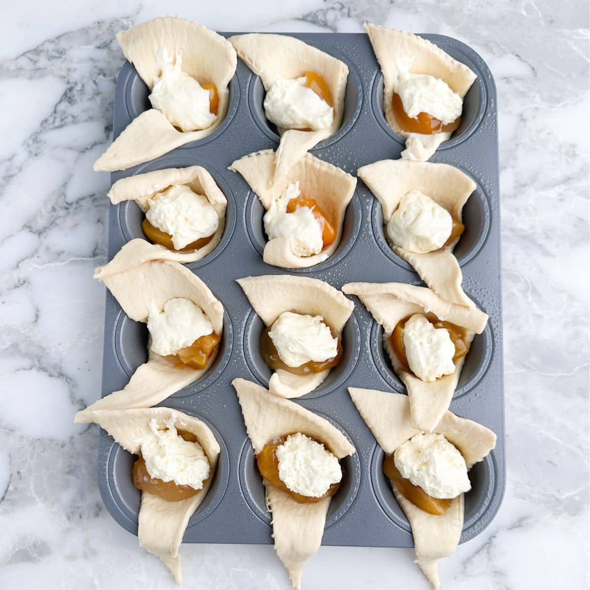 Muffin tin with uncooked crescent rolls, peach pie filling, and a dollop of cheesecake filling.