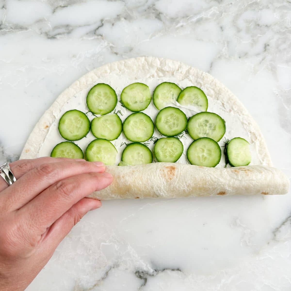 Tortilla with cream cheese and sliced cucumbers being rolled up. 
