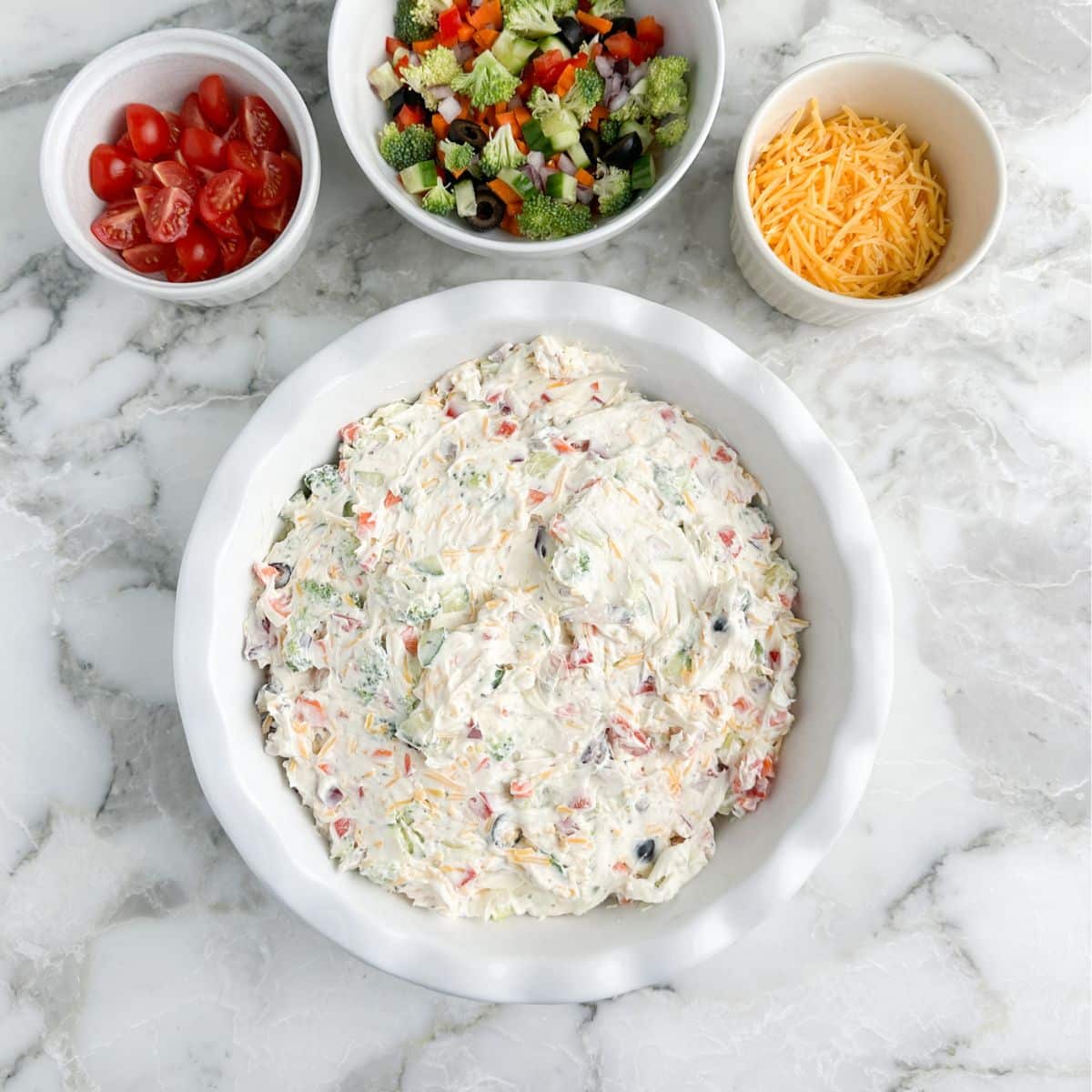 Baking dish with vegetable dip, bowl of chopped vegetables, tomatoes, and shredded cheese.