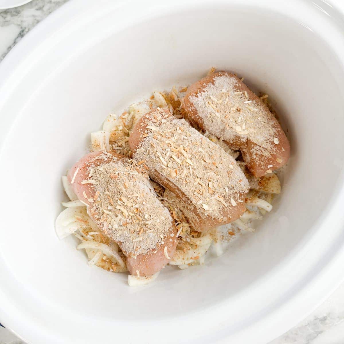 Slow cooker with onions, chicken breasts, and seasonings.