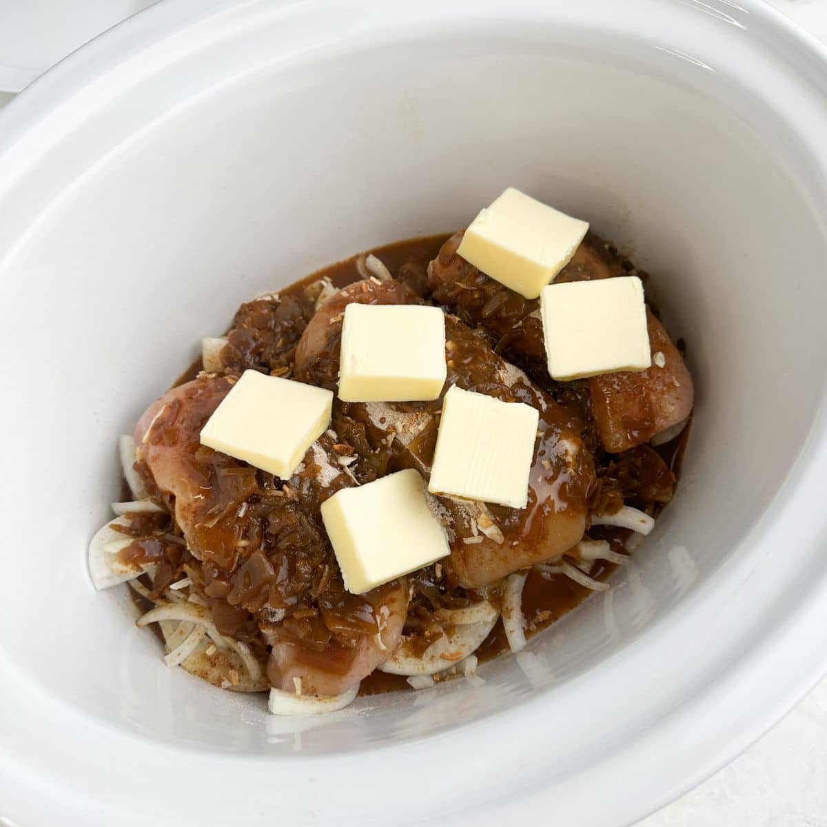 Slow cooker with onions, chicken, seasonings, and slices of butter.