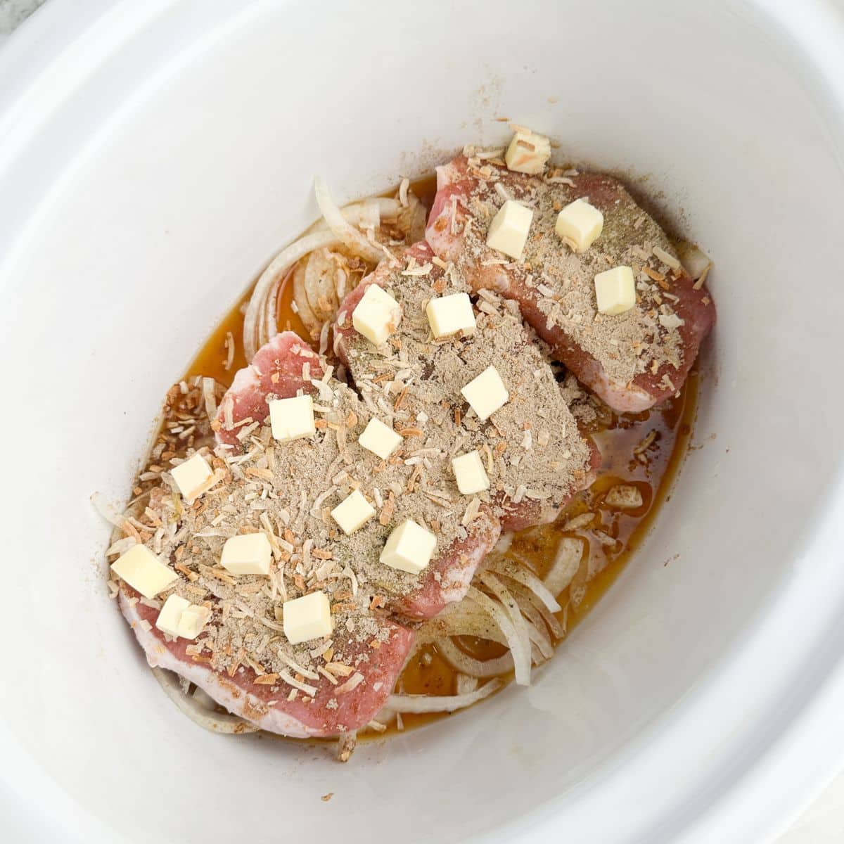 Slow cooker with onions, pork chops, onion soup mix, and pats of butter.