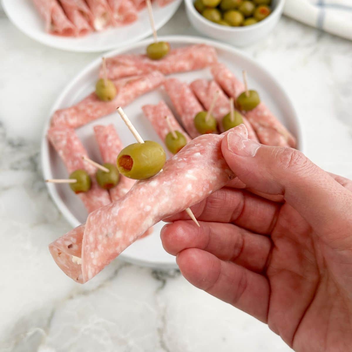 Hand holding a salami roll up with a green olive.