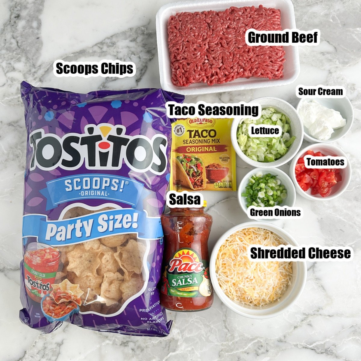 Bag of chips, ground beef, taco seasonings, salsa, shredded cheese, bowls of lettuce, tomato, sour cream, and green onions.