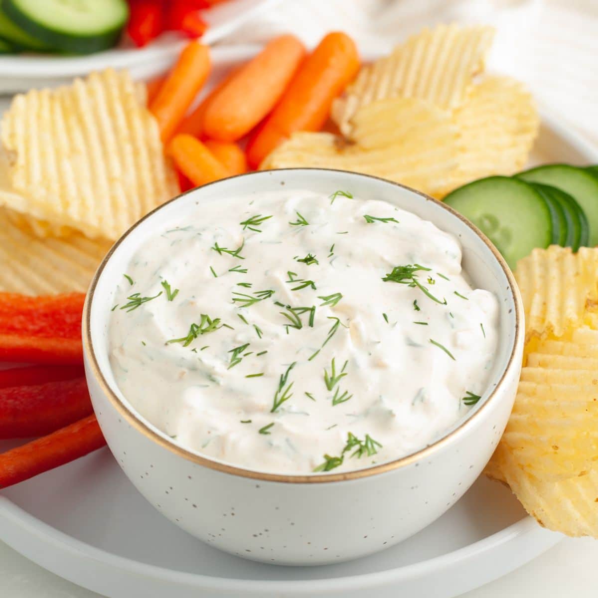 Bowl of dill dip surrounded by potato chips and vegetables. 
