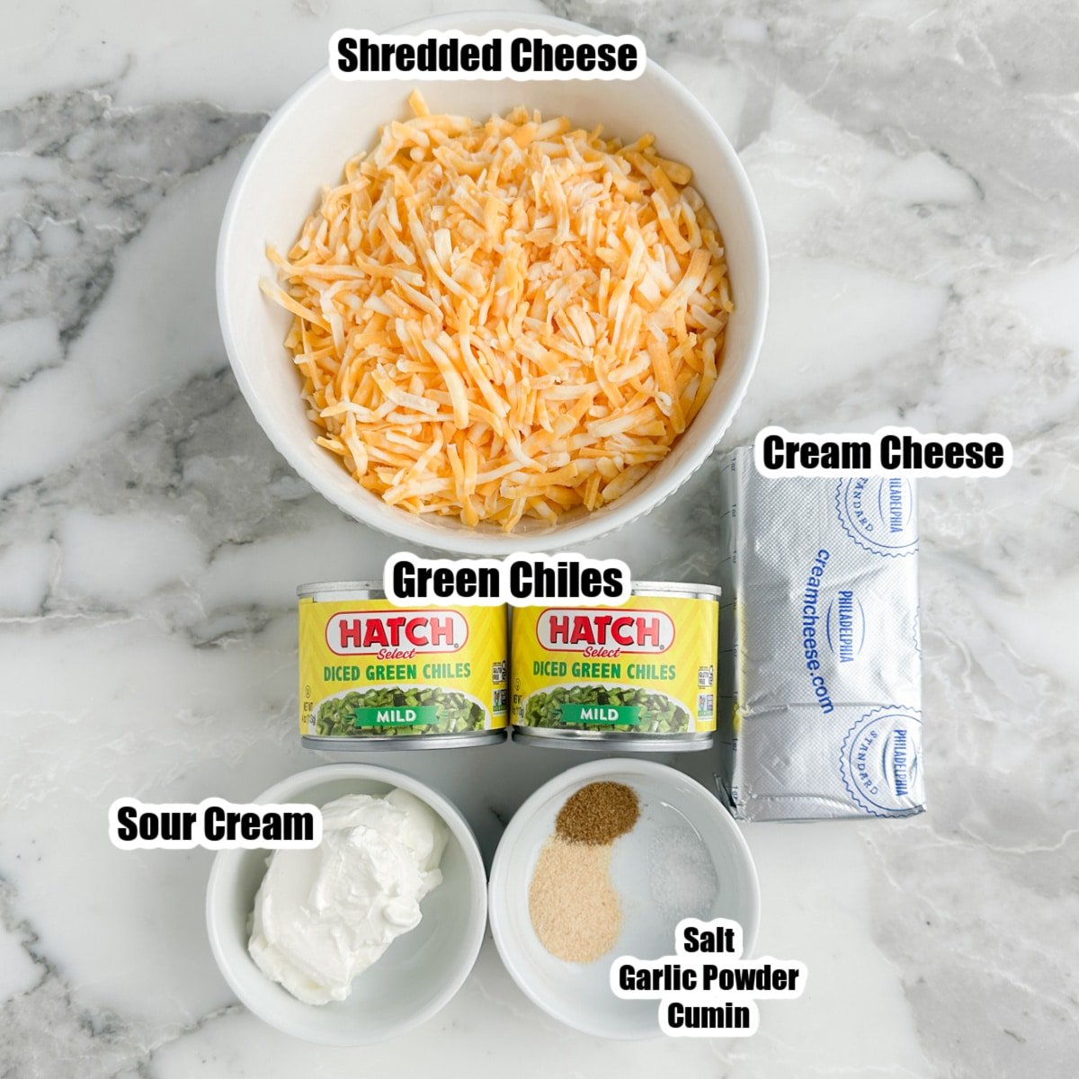 Bowl of shredded cheese, sour cream, cream cheese, cans of green chiles, bowl of salt, garlic powder, and cumin.