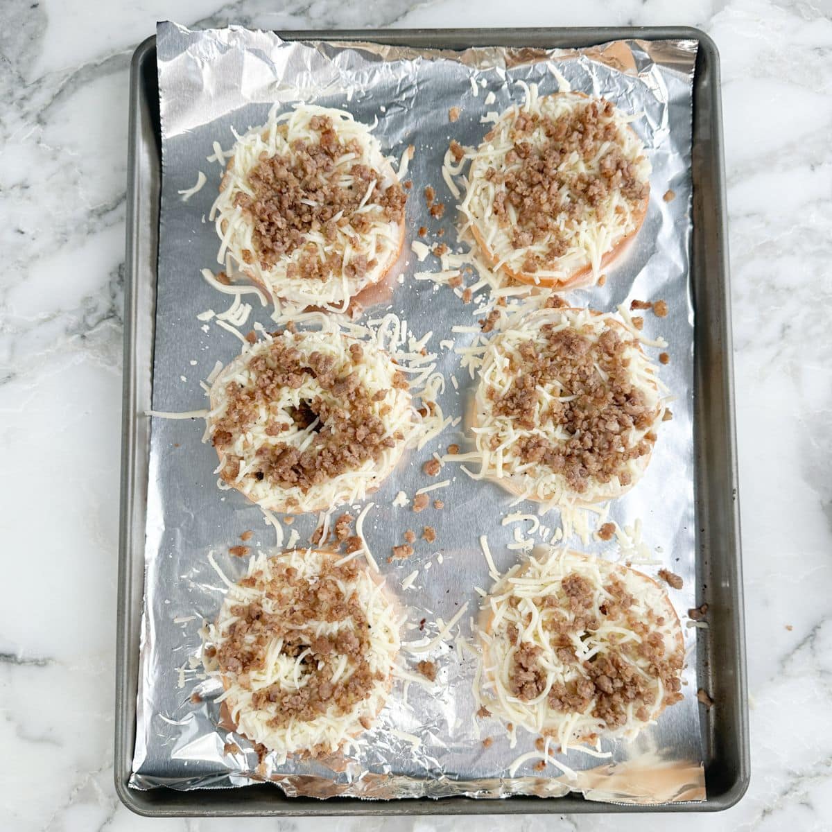 Baking pan with bagels topped with cheese and crumbled sausage.