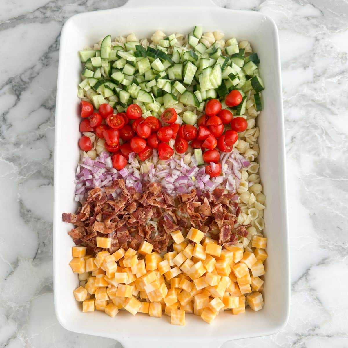 Casserole dish with pasta, diced cucumbers, tomatoes, onion, bacon crumbles, and cubed cheese. 