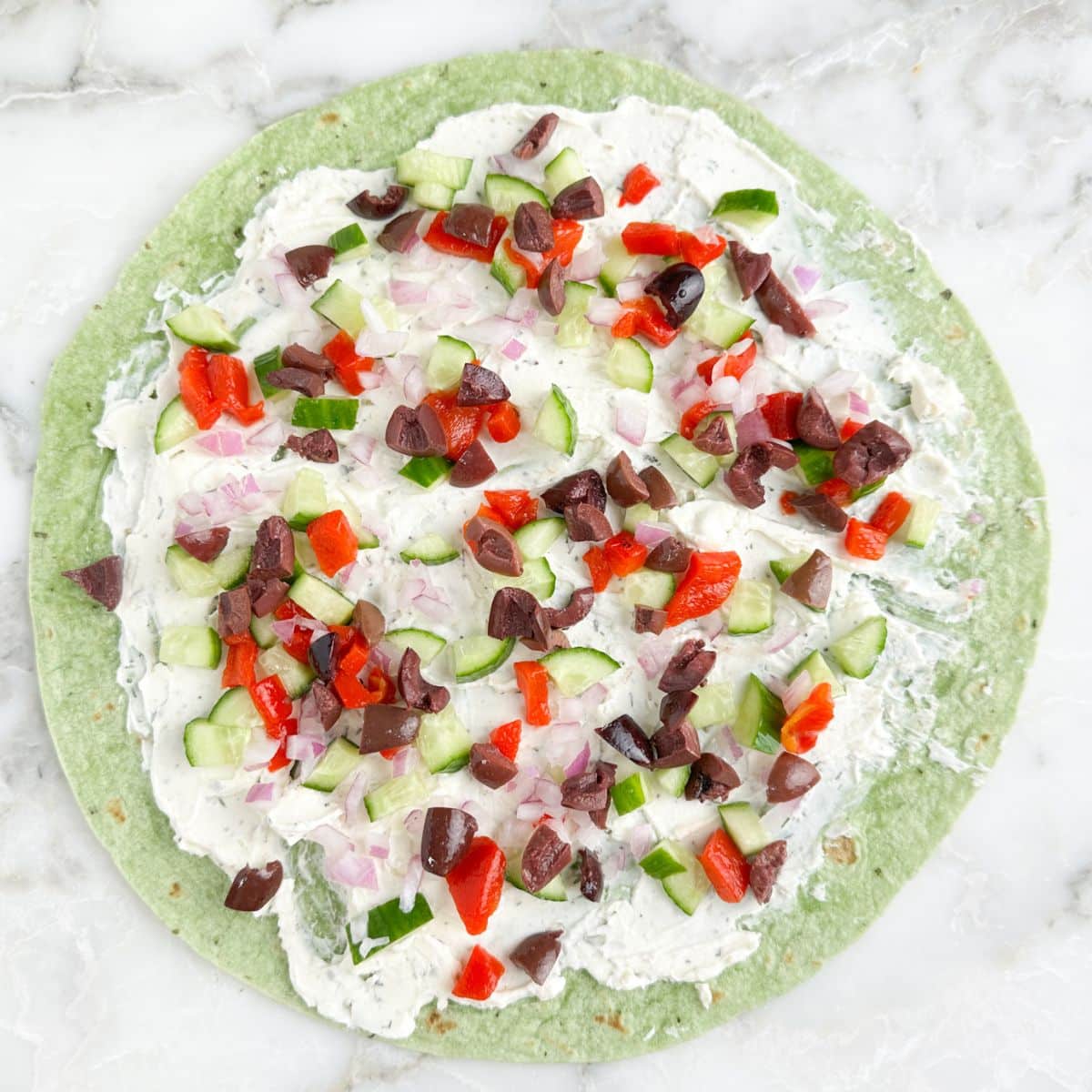 Green tortilla topped with cream cheese spread, cucumber, peppers, and olives.