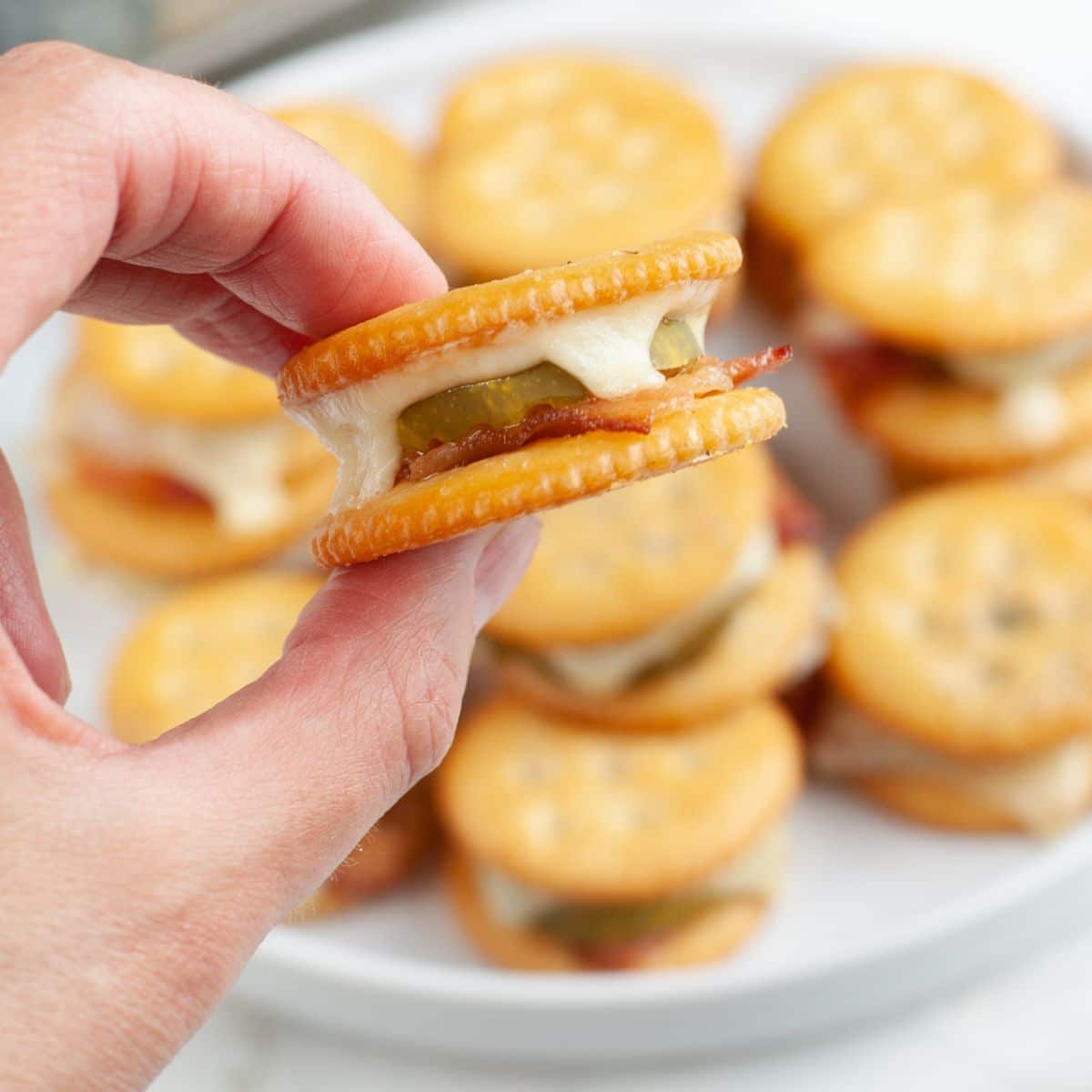 Hand holding Ritz cracker with bacon and pickle.