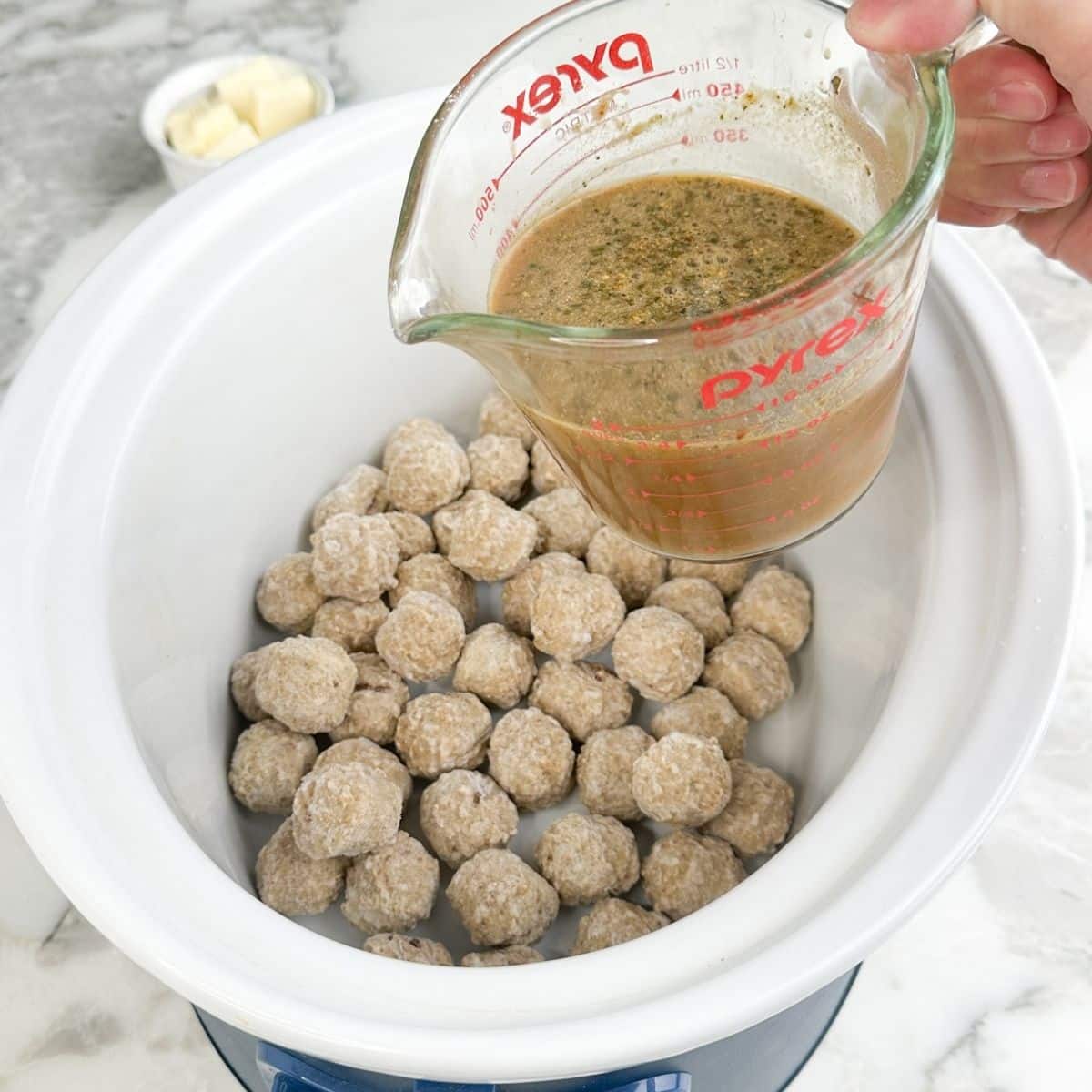 Frozen meatballs in a slow cooker with broth in a measuring cup.